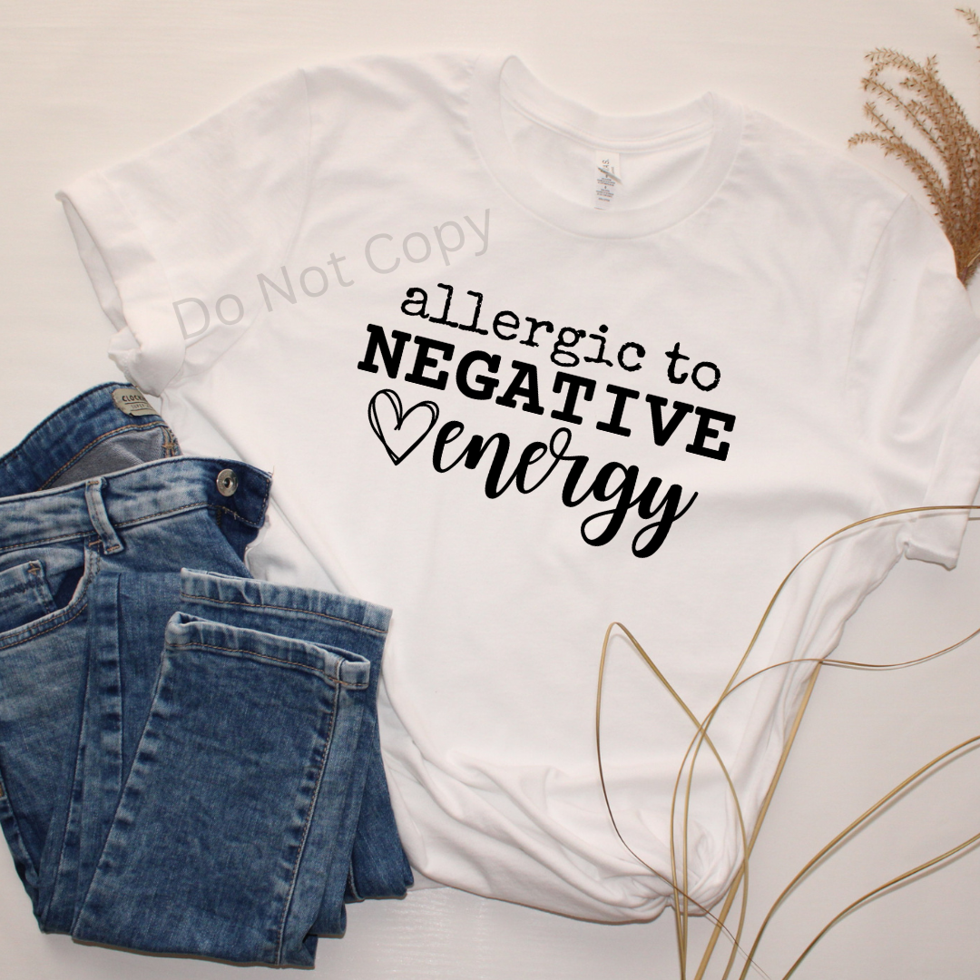 A white crew neck t-shirt with the phrase 'allergic to negative energy' printed in black, accompanied by a heart symbol. The image is presented alongside a pair of blue jeans and dry wheat, indicating a casual clothing style.