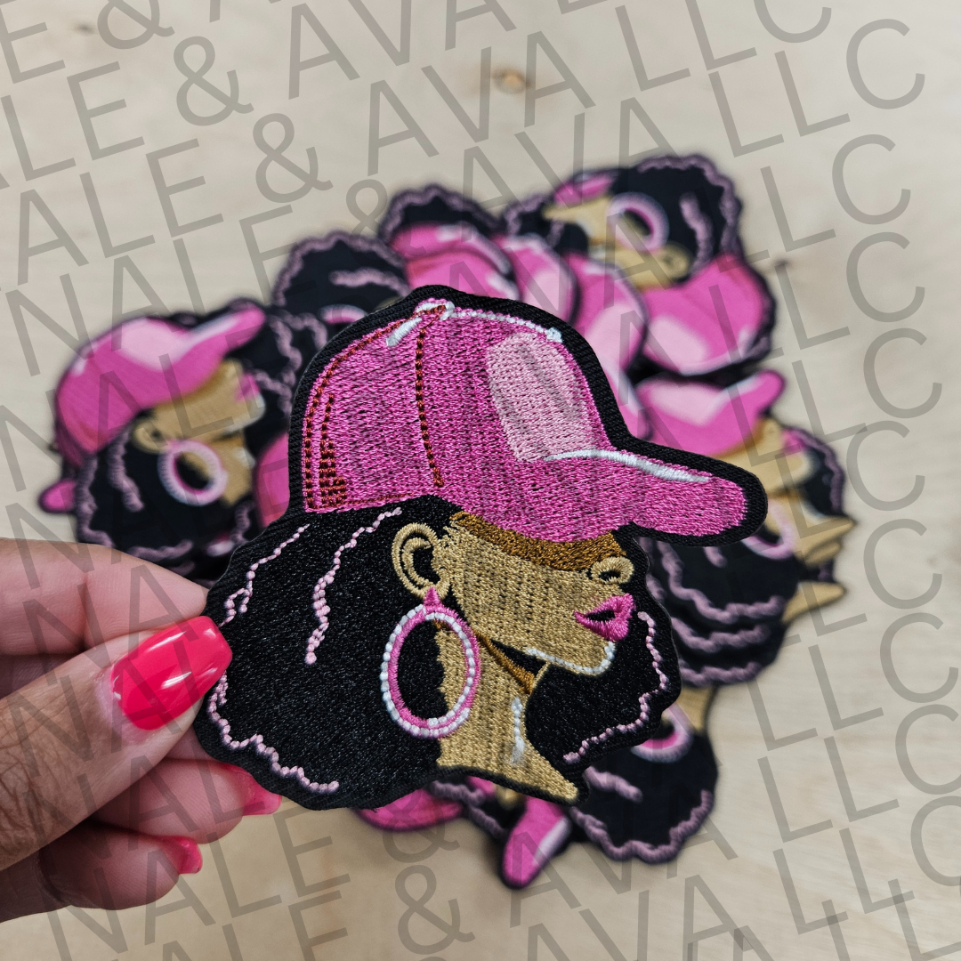 Embroidered patch of a person wearing a pink cap held by a hand on a patterned background.
