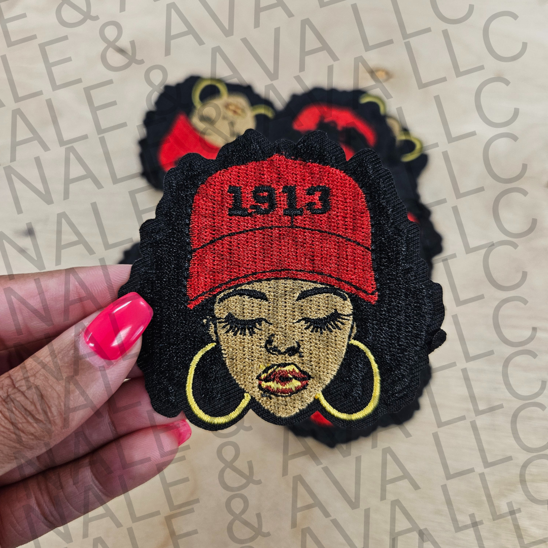 Embroidered patch of a woman wearing a red '1913' cap held by a hand on a branded background.