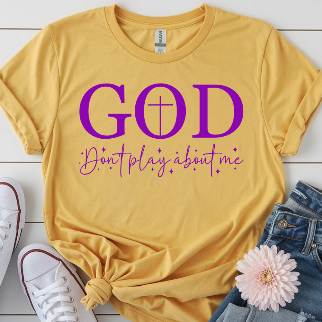 Yellow t-shirt with purple text 'GOD Don't play about me' on a wooden surface with shoes and jeans.