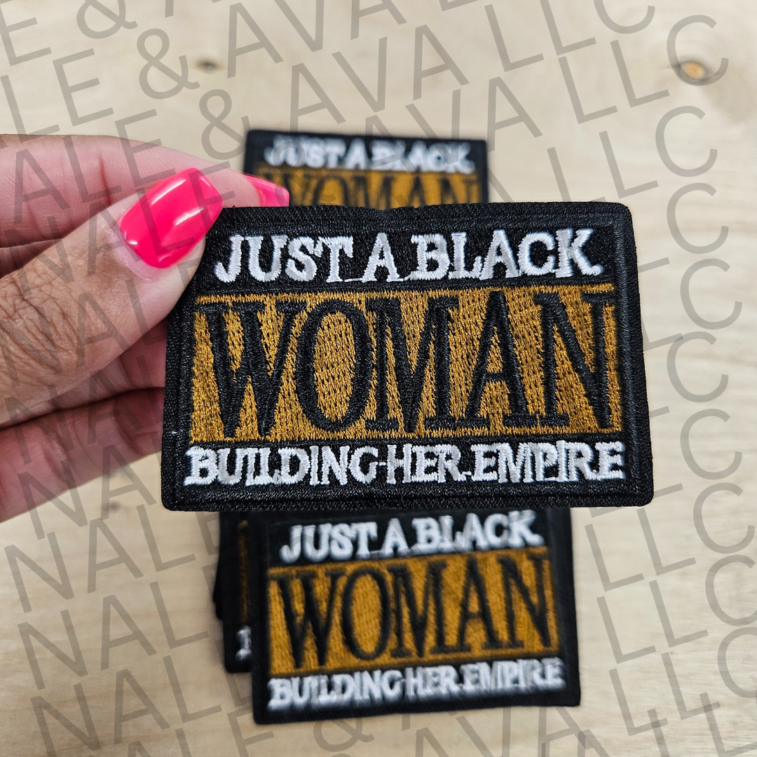 Patch with 'Just a Black Woman Building Her Empire' text held by a hand with pink nail polish on a beige background.