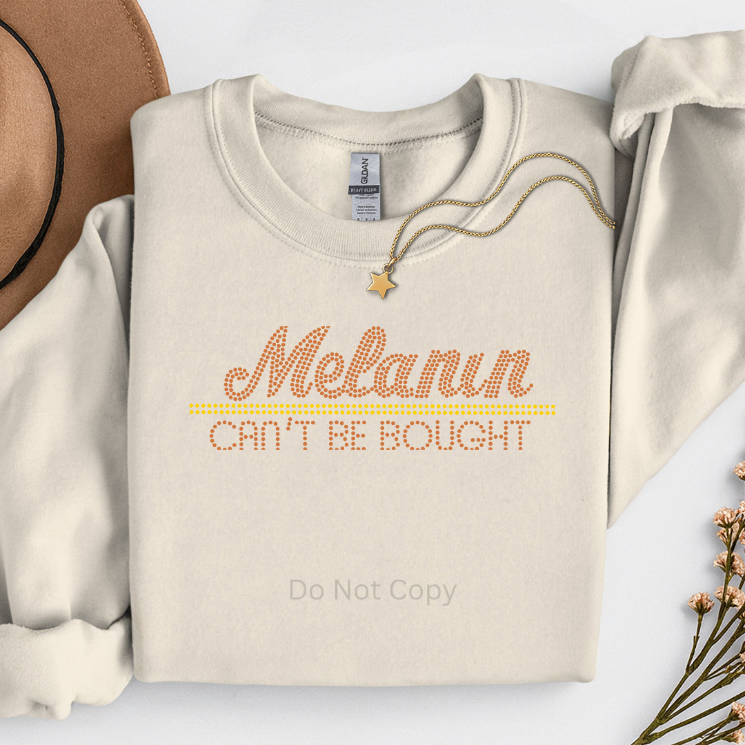 Beige sweatshirt with 'Melanin Can't Be Bought' text, surrounded by a hat and dried flowers on a light background.