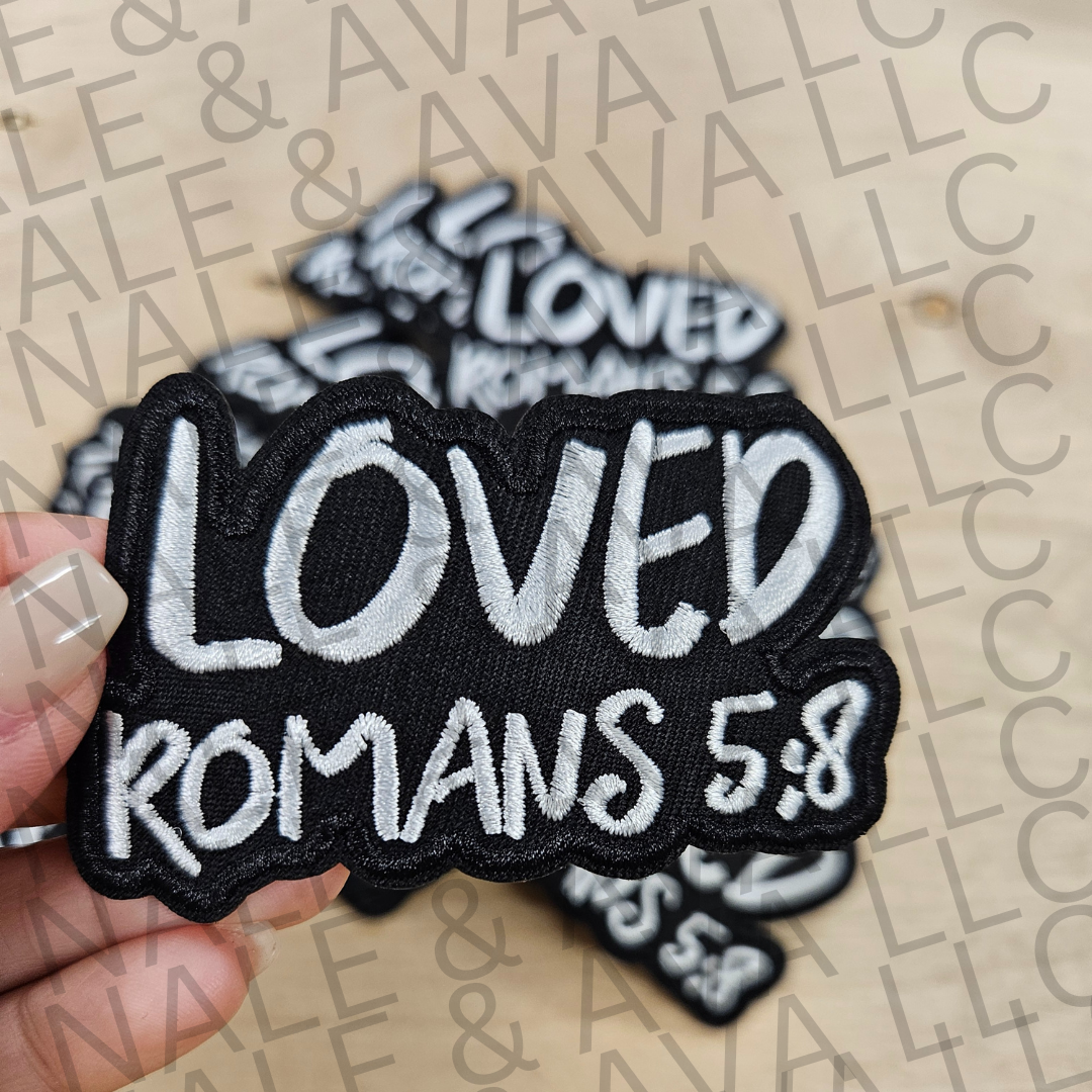 Black and white 'LOVED ROMANS 5:8' patch held by a hand on a beige background with 'Avalon' branding.