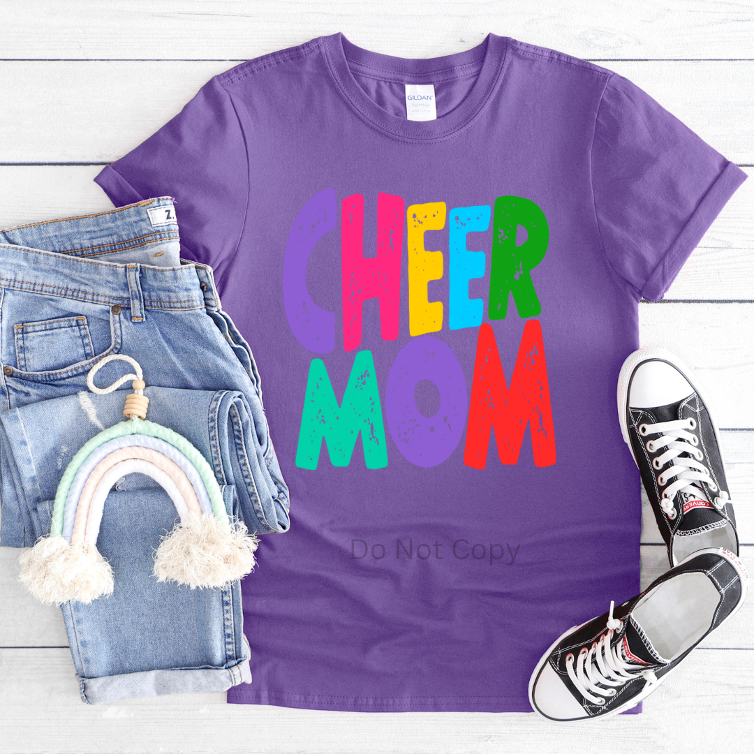 A purple t-shirt with the text 'CHEER MOM' in colorful letters, accompanied by a pair of jeans and a pair of sneakers, presented on a wooden surface.