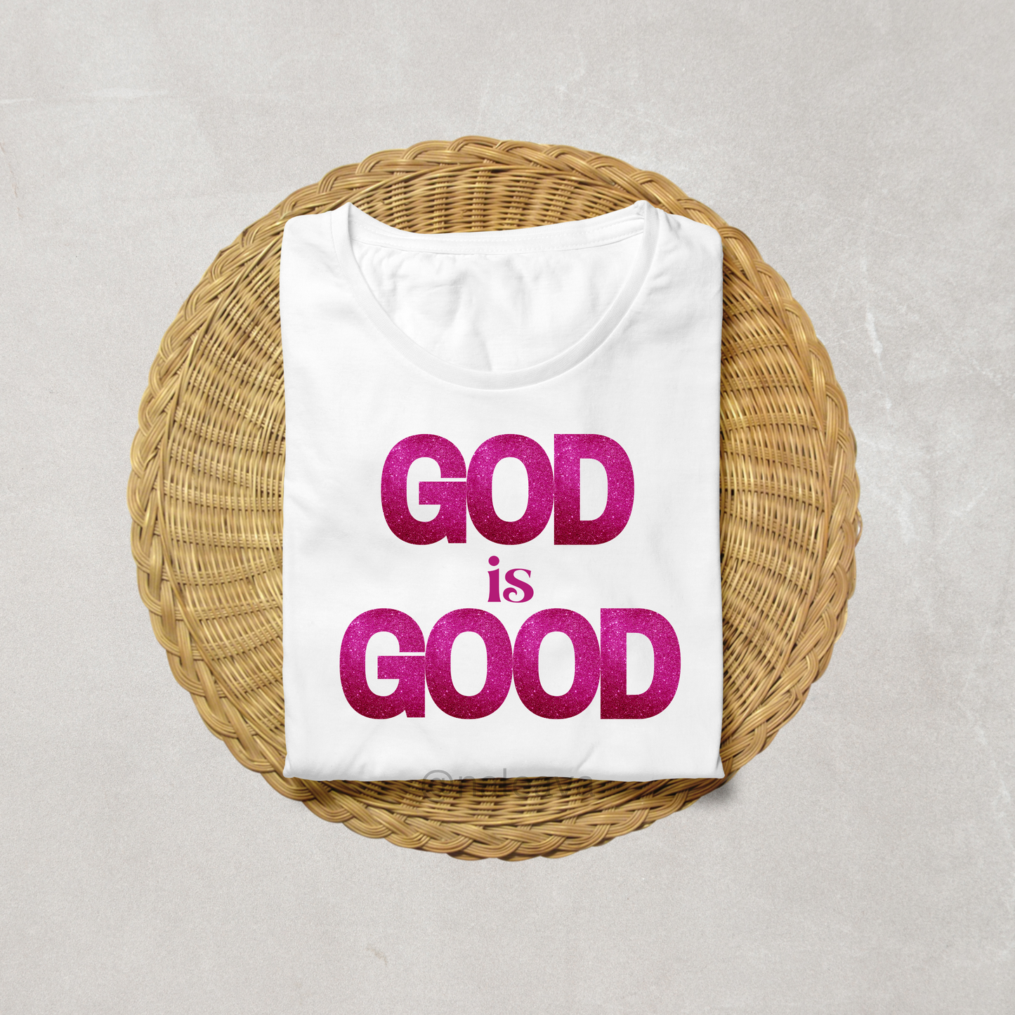 A white T-shirt with the text 'GOD IS GOOD' printed in bold pink letters on the front, placed on a woven basket on a textured background.