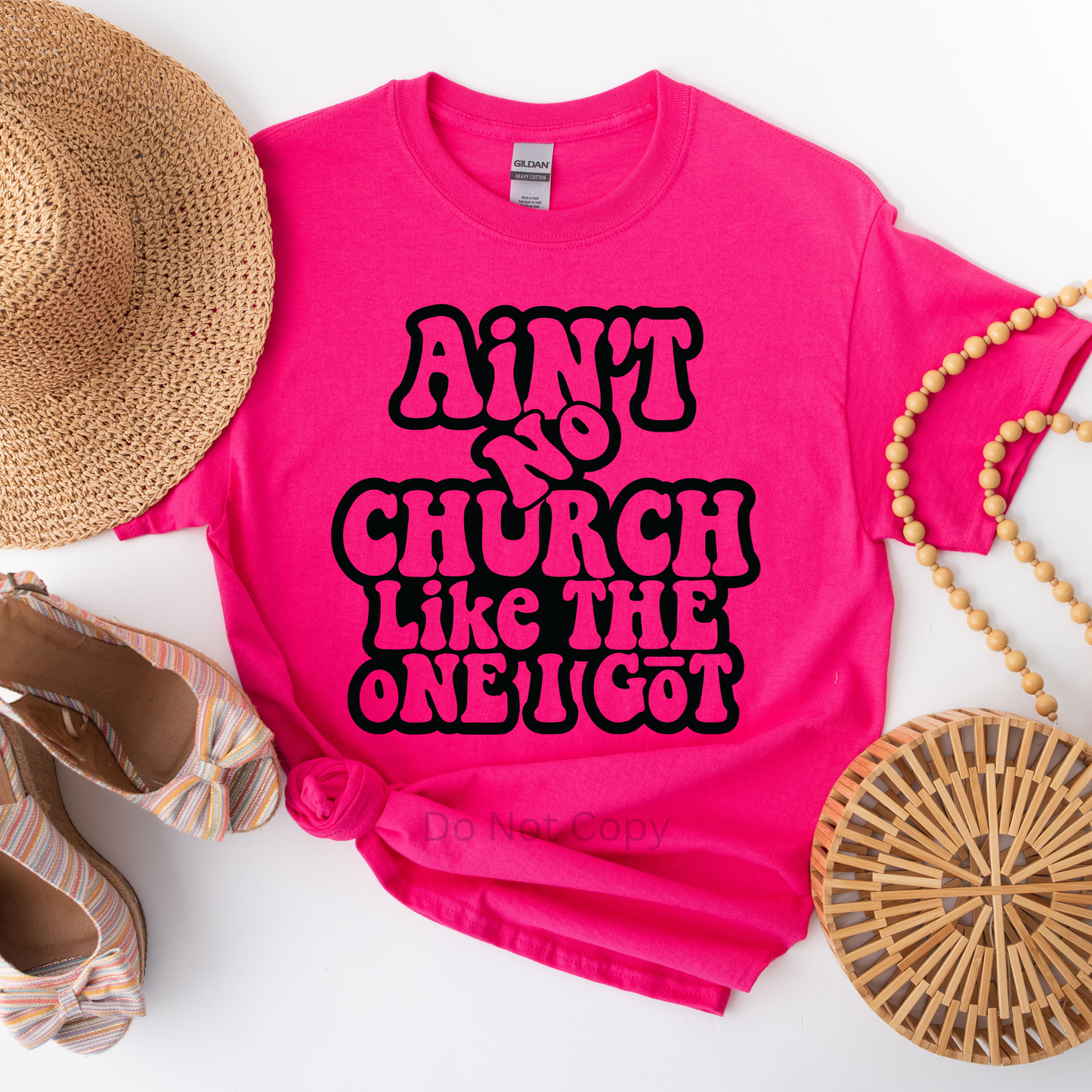 A pink T-shirt with the text 'AIN'T NO CHURCH LIKE THE ONE I GOT' in bold black letters, displayed alongside a straw hat, pearls, and patterned shoes.