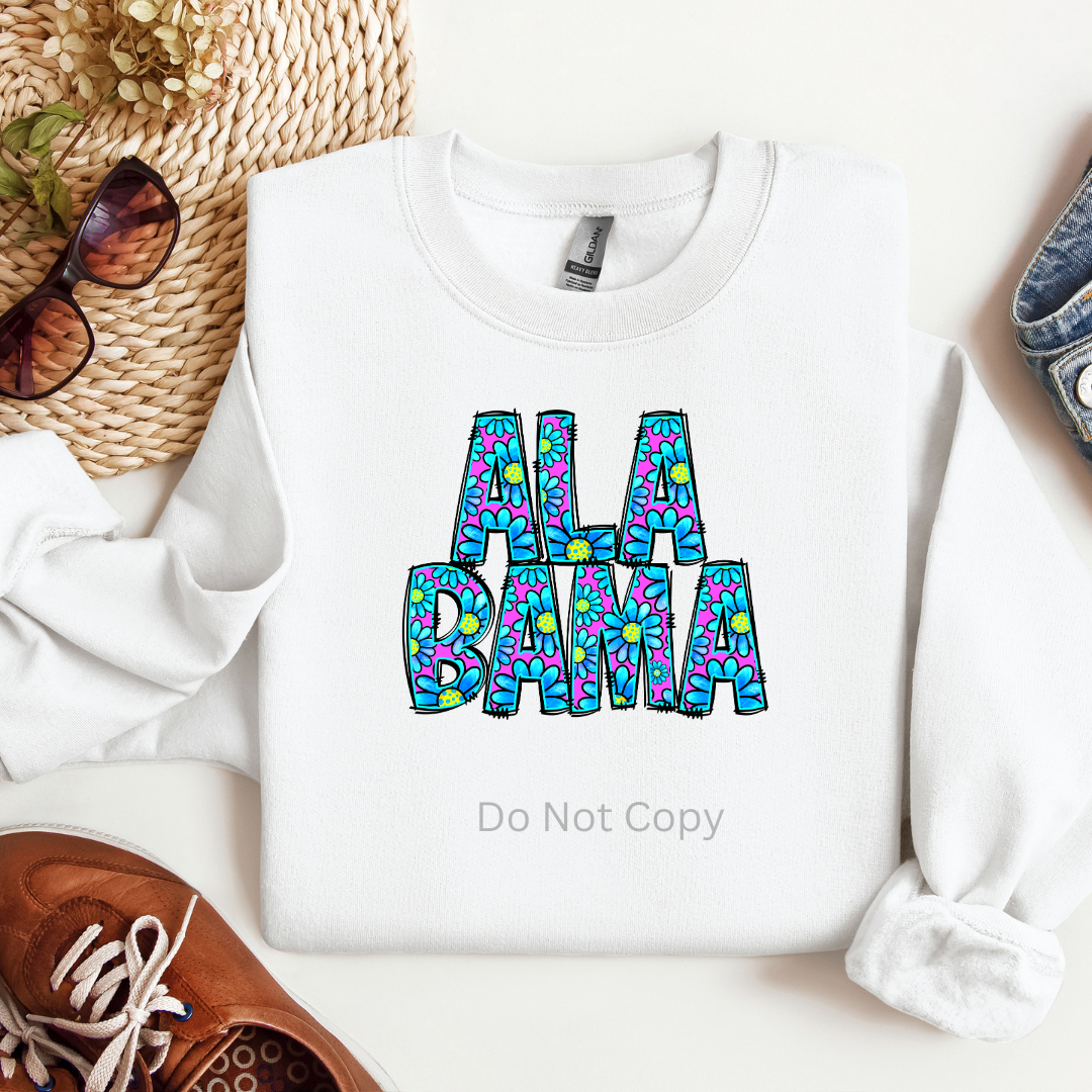 A white garment with the text 'ALABAMA' printed on it in a stylized font, accompanied by sunglasses, a woven hat, and a pair of shoes on a white background.