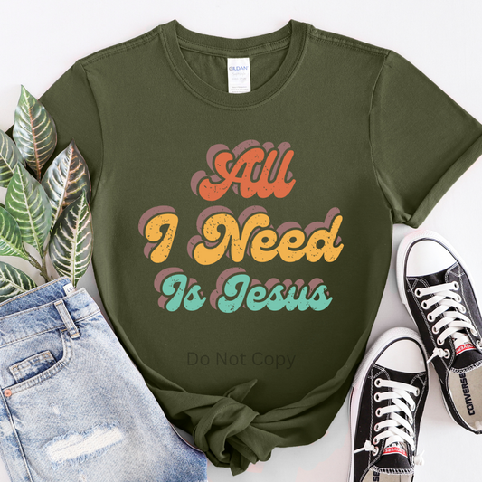 A green T-shirt with the text 'All I Need Is Jesus' printed in a multicolor font, displayed alongside denim jeans and black sneakers.