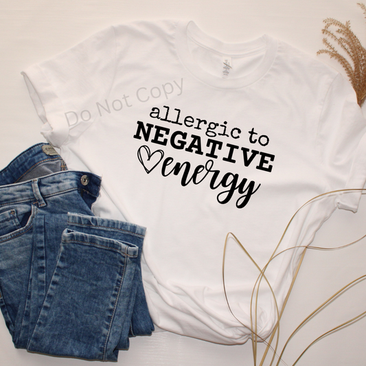A white crew neck t-shirt with the phrase 'allergic to negative energy' printed in black, accompanied by a heart symbol. The image is presented alongside a pair of blue jeans and dry wheat, indicating a casual clothing style.