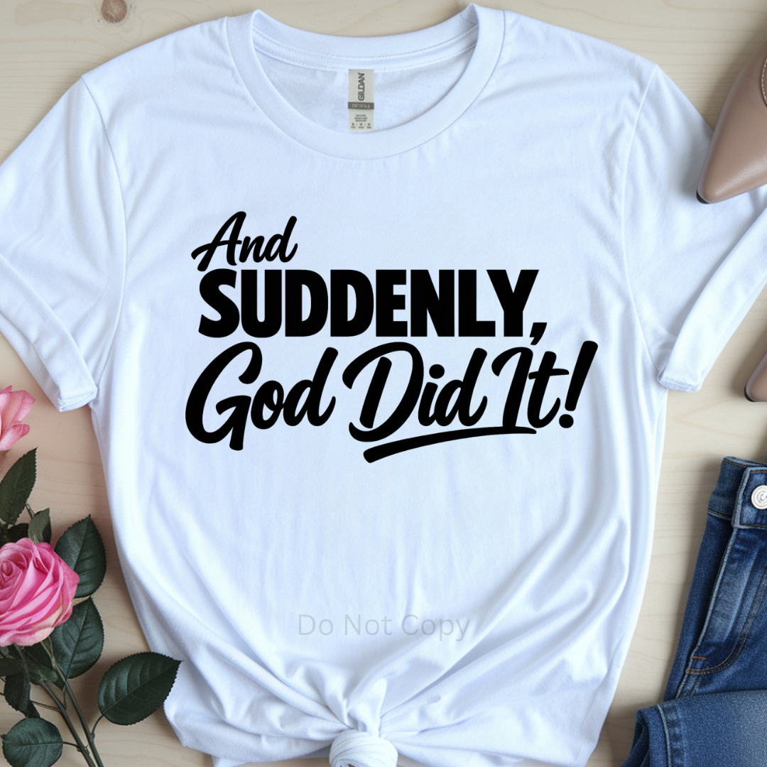 White t-shirt with black text 'And Suddenly, God Did It!' on a light wooden surface with flowers and jeans.