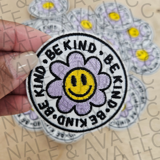 Patch with a smiley flower design and 'Be Kind' text held by a hand, with more patches in the background.