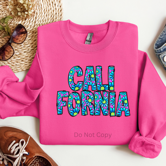 A pink sweatshirt with the text 'CALIFORNIA' printed on it in a stylized font, surrounded by a pattern of what appears to be leaves or flowers in various colors.
