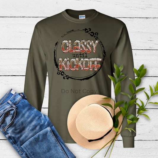 A long-sleeved green T-shirt with a graphic transfer design that reads 'CLASSY until KICKOFF' surrounded by decorative elements, displayed alongside a pair of blue jeans and a straw hat, against a white wooden background.