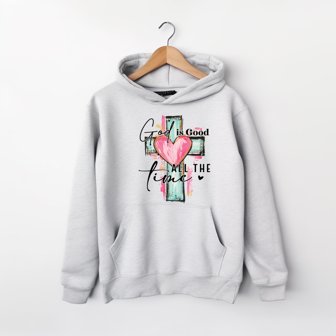 A light gray pullover hoodie hanging on a wooden hanger against a white wall. The hoodie features a front graphic with a pink heart, a teal and pink cross, and the text 'God is Good ALL THE time' in black script and block lettering.
