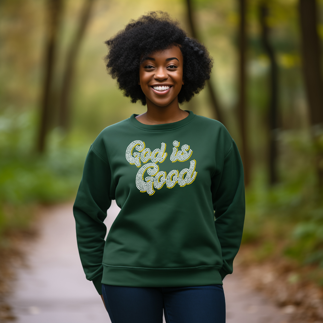 A smiling woman with an afro stands on a forest path wearing a dark green crewneck sweatshirt with 'God is Good' written across the chest in a glittery gold and silver script.