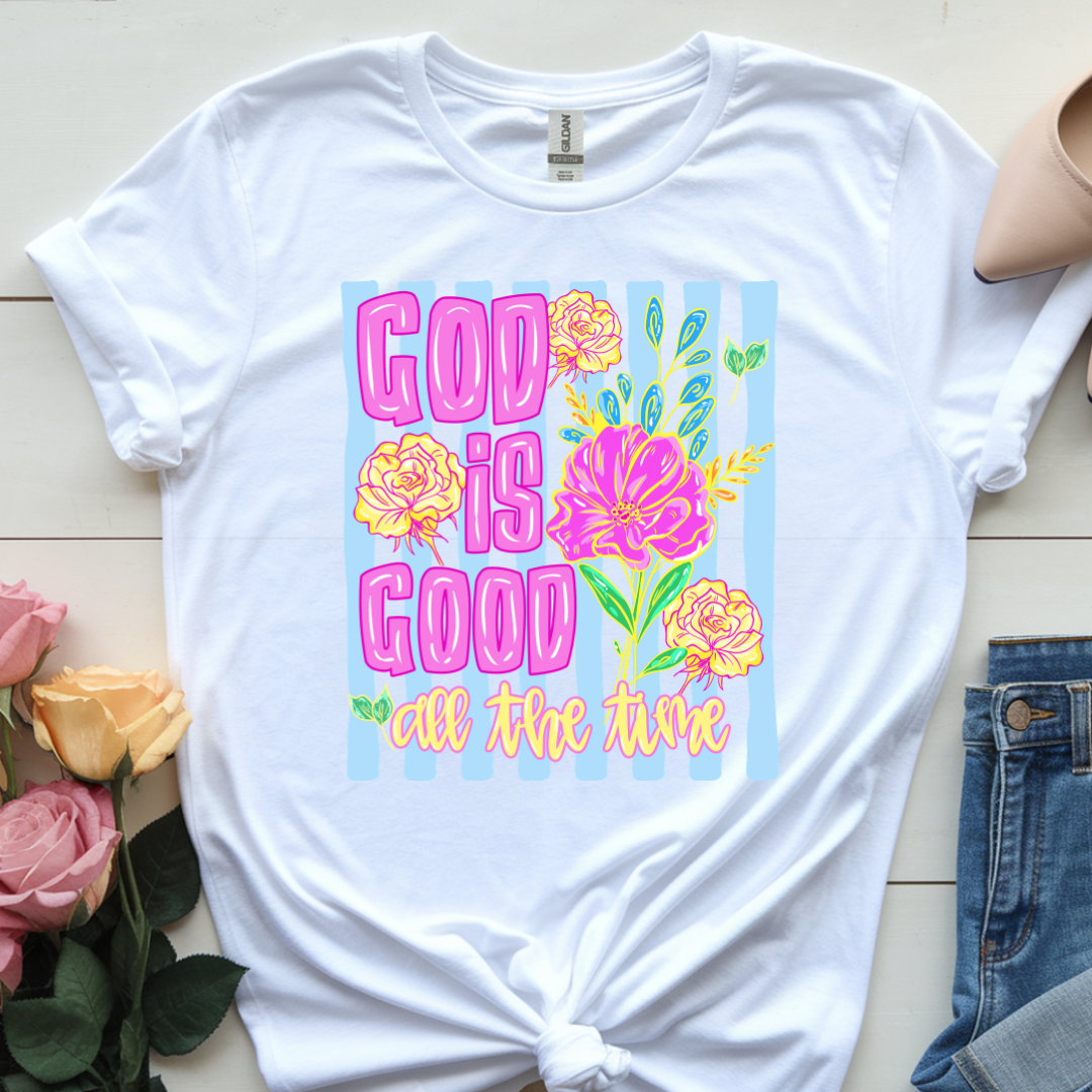 White t-shirt with 'God is Good all the time' text and floral design on a wooden surface with flowers and jeans.
