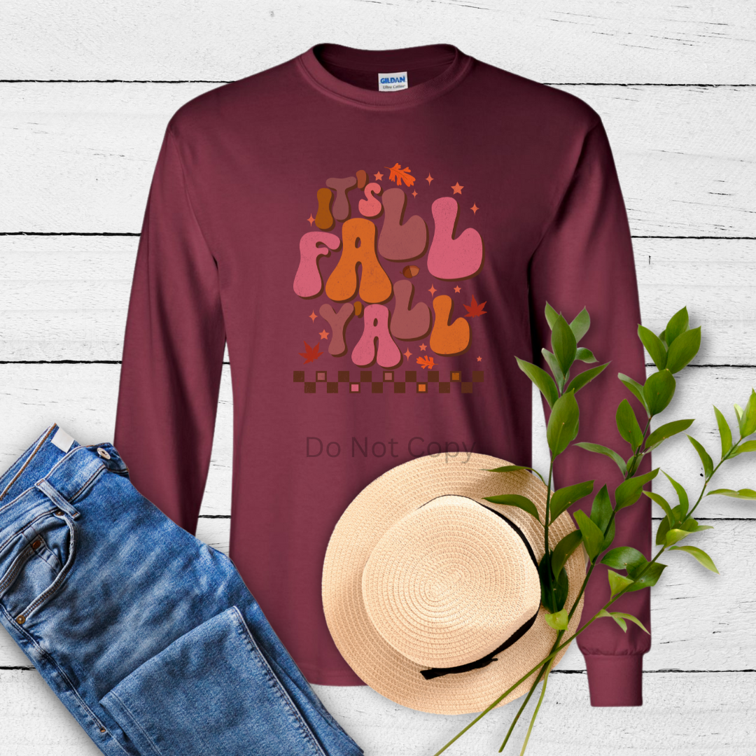 A maroon long-sleeve shirt with a text and colorful design on the front, accompanied by a blue jean, a straw hat, and a heat press, displayed on a wooden surface with green leaves in the background.