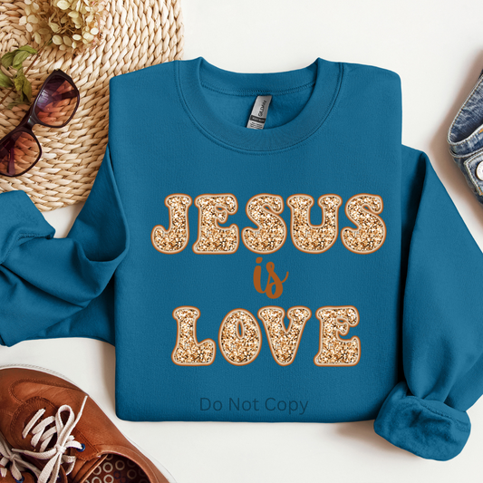 A blue sweatshirt with the text 'JESUS IS LOVE' and a sparkle effect on the letters, displayed on a flat surface with sunglasses and a shoe beside it.