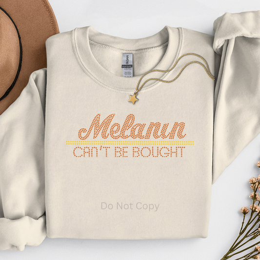 Beige sweatshirt with 'Melanin Can't Be Bought' text, surrounded by a hat and dried flowers on a light background.
