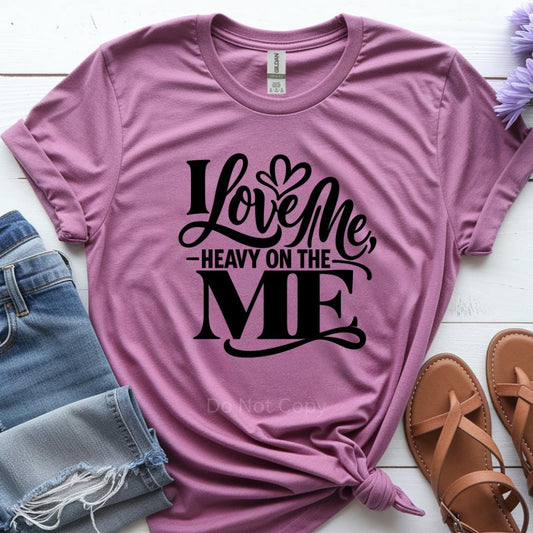 Pink t-shirt with 'I Love Me, Heavy on the ME' text, paired with denim shorts and sandals on a white surface.