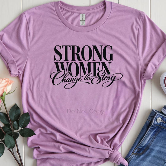 Purple t-shirt with 'Strong Women Change the Story' text on a white surface with flowers and jeans.