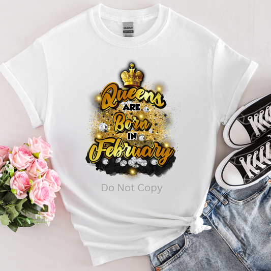 A white T-shirt with the text 'Queens are Born in February' printed on it, accompanied by a crown and decorative elements, placed next to a pair of sneakers and a bouquet of flowers.