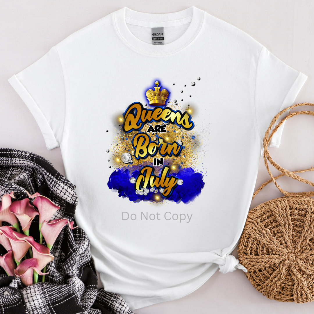 A white T-shirt with the text 'Queens are Born in July' printed on it, featuring a crown and decorative elements in gold and blue.
