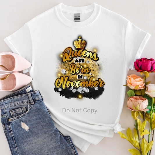 A white T-shirt with the text 'Queens are born in November' printed on it, accompanied by flowers and a pair of shoes.