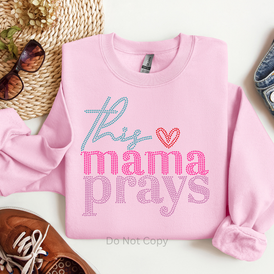 Pink sweatshirt with 'This mama prays' text on a white surface with accessories.