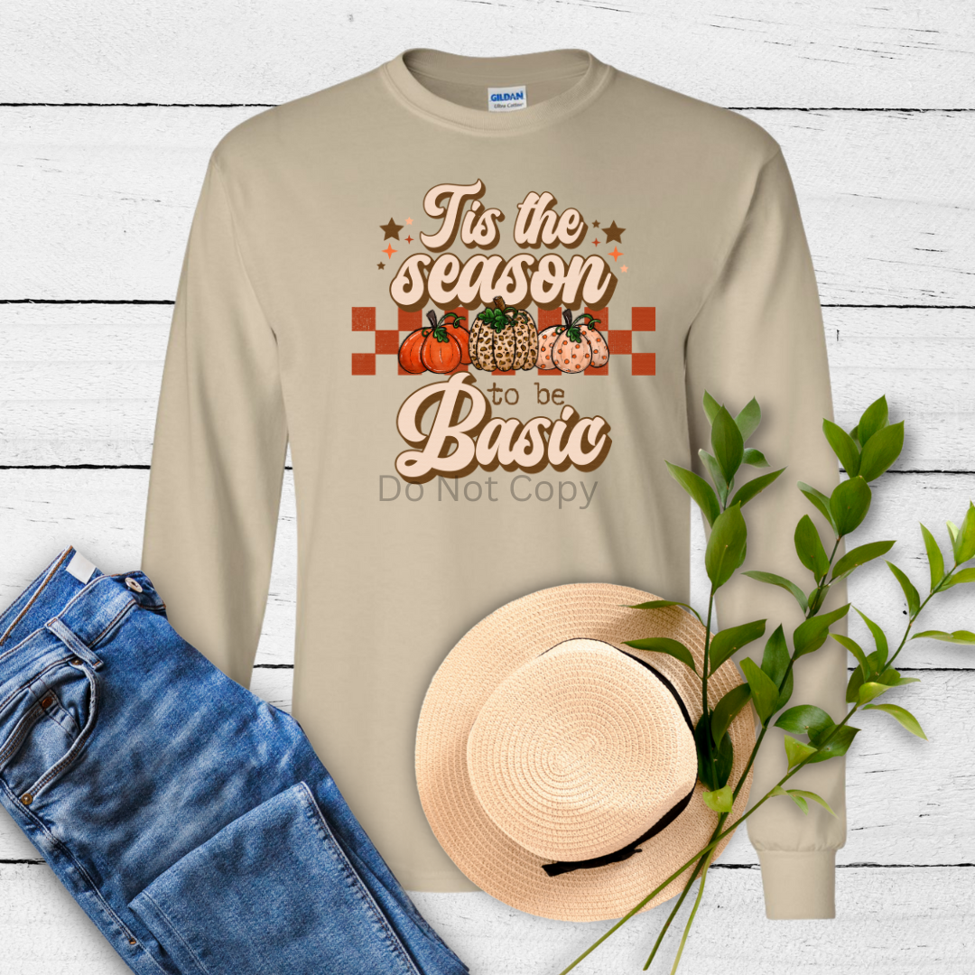 A beige long-sleeve shirt with a printed text 'It's the Season to be Basic' accompanied by fall-themed images like pumpkins and apples on the front. A hat and jeans are also visible in the background, suggesting a casual, seasonal themed display.