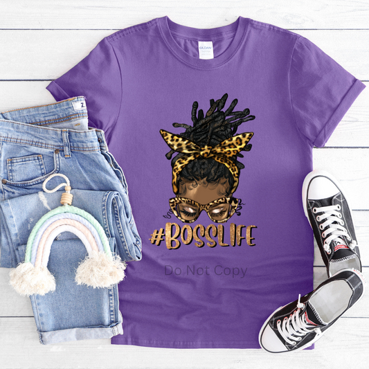 A purple T-shirt featuring a digital print of a woman's character with accessories and the text '#BossLife'. The shirt is displayed flat alongside a fabric roller, jeans, and sneakers.