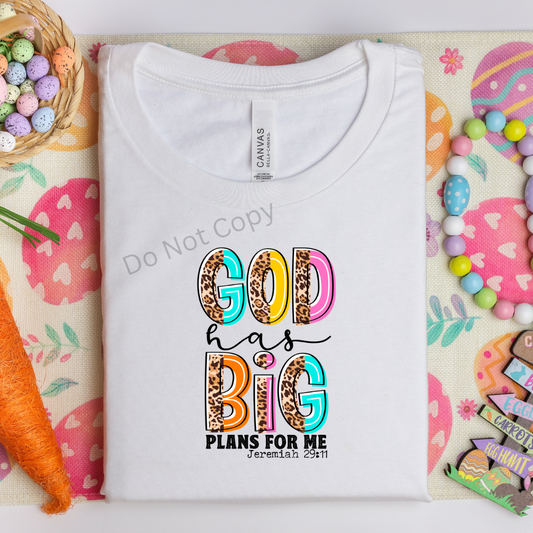 A white t-shirt with the text 'GOD HAS BIG PLANS FOR ME' printed on it, laid flat on a surface with decorative elements around it.