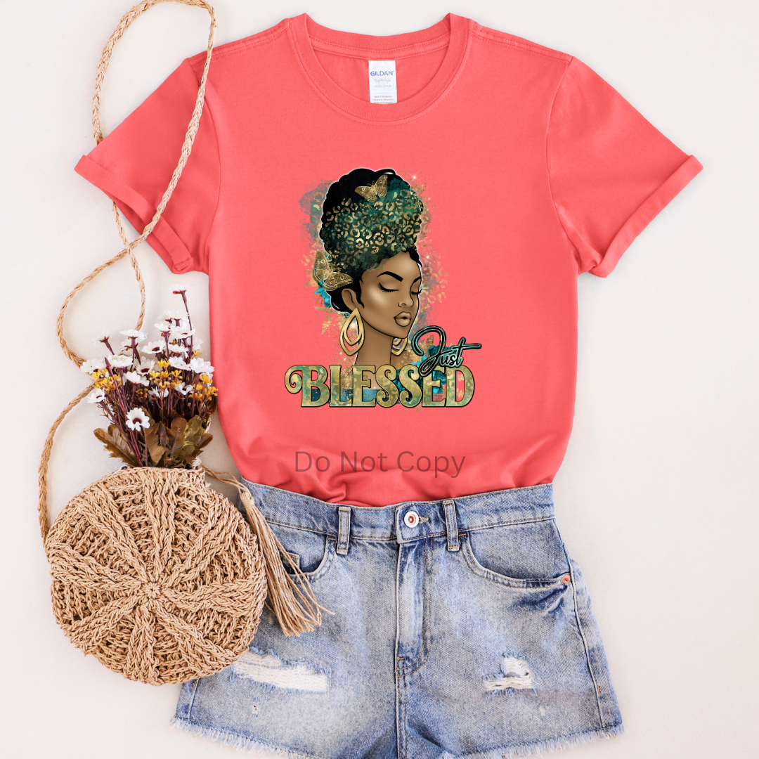 A red T-shirt featuring a printed transfer with a woman's image and the text 'Blessed'. The shirt is displayed alongside a beige fabric roller and a straw tote.