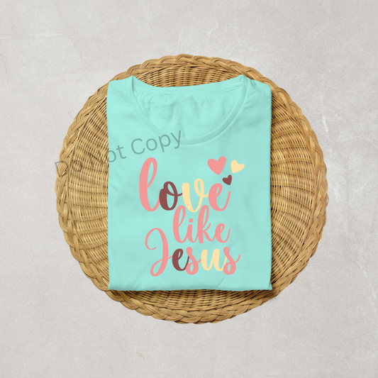A folded light blue t-shirt with the text 'love like Jesus' printed on it, placed on a woven mat.